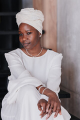 Elegant African woman in a white dress and headscarf sitting thoughtfully. Brazilian girl relaxing...
