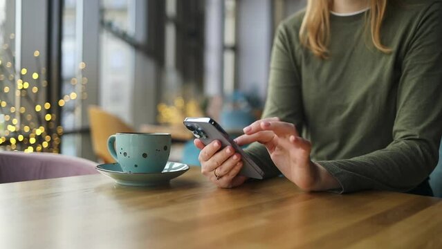 Crop view of female person holding smartphone and scrolling mobile phone while sitting at table with coffee at coffee shop cafe. Work people contact marketing business, technology. High quality FullHD