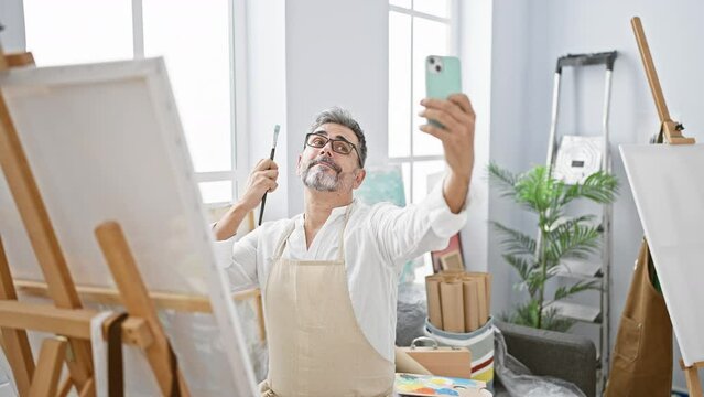 Smiling young, grey-haired hispanic man artist captures a joyful selfie with smartphone amidst paintbrushes and canvas in art studio