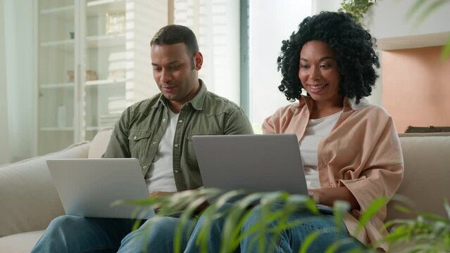 Multiracial couple in love working on laptops at home African American family business people work together on couch shopping online on computers looking at camera smiling woman and man use devices