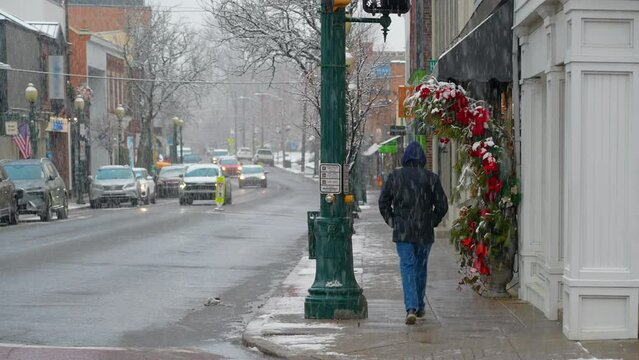A man walks away from the camera on the sidewalk of a small New England town on a snowy winter day during the Christmas season. Pittsburgh suburbs.	