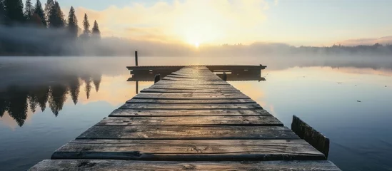 Fotobehang A wooden pier stretches into the calm lake, with fog above. The tranquil water mirrors the pier and the sunrise creates a beautiful sky. © TheWaterMeloonProjec