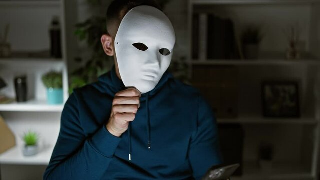 A young hispanic man in a hoodie holding a smartphone and a white mask in a modern office setting