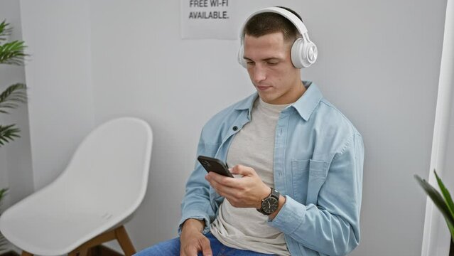 Hispanic man in headphones uses smartphone in a modern waiting room, exuding a casual, tech-friendly vibe.