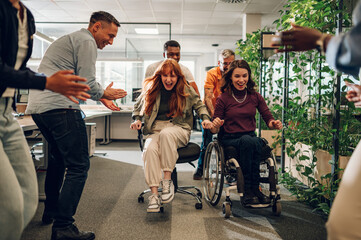Woman who uses a wheelchair and female colleague racing on chairs in an office