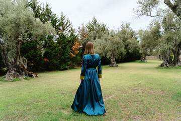 A woman in a teal dress stands contemplative among ancient olive trees in a peaceful grove