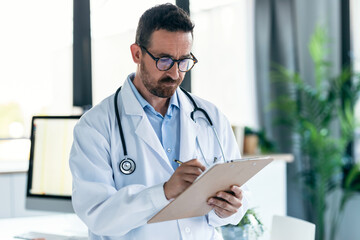 Mature male doctor with stethoscope over neck taking notes in clipboard in a medical consultation