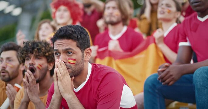 Group of multi-ethnic fans sitting on stadium tribunes and showing their disappointment. Sad from negative outcome. Supporting their national Spanish team. Wearing Spanish flags painted on their faces