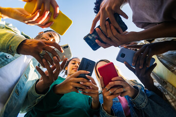 Diverse group of young people using mobile phone device standing together in circle outdoors. Millennial friends addicted to social media app, betting or playing video game on platform online. - obrazy, fototapety, plakaty