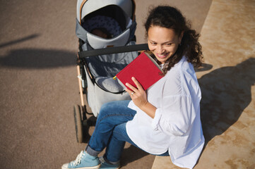 View from above of authentic delightful young mother smiling looking aside, enjoying reading book...
