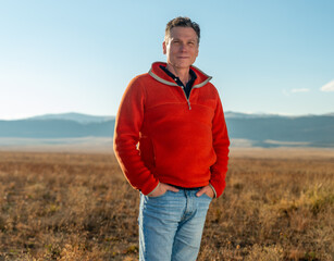 Middle aged man farmer, rancher, real estate developer, CEO, in orange fleece pullover standing on vast high plains grassland with Rocky Mountains in the background