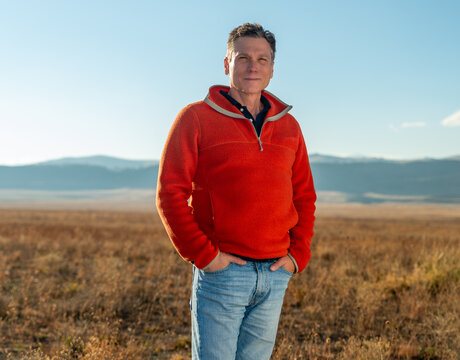 Middle aged man farmer, rancher, real estate developer, CEO, in orange fleece pullover standing on vast high plains grassland with Rocky Mountains in the background - Powered by Adobe