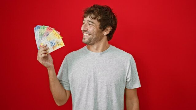 Happy young man with blond beard proudly holding up swiss franc banknotes, financial triumph writ large on his face, depicted in an isolated, vivacious red background portrait!