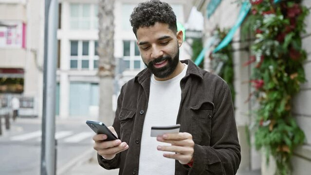A smiling young adult man with a beard looks at his phone while holding a credit card on a busy urban street.
