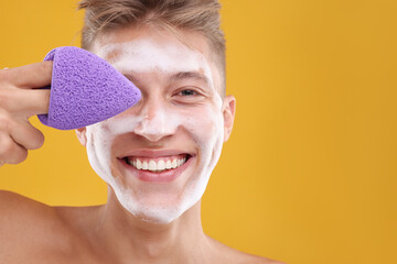 Happy young man washing his face with sponge on orange background. Space for text