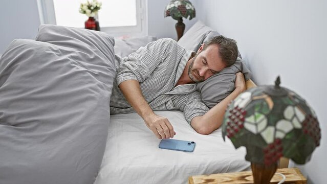 A middle-aged man reaching for a smartphone in bed, depicting an everyday indoor scene of modern life.