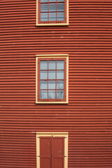 The exterior of a red colored cottage with wooden wall covered in horizontal clapboard siding. There are two vintage windows with multiple glass panes. The decorative trim is yellow and orange colored