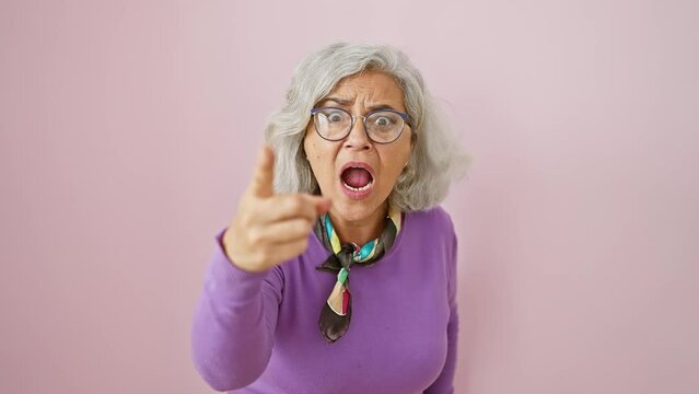 Furious middle-aged woman, grey-haired and bespectacled, pointing accusing finger at you in front of isolated pink background, oozing anger and frustration, clearly displeased.