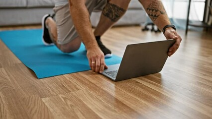 A man with tattoos sets up a laptop on a yoga mat for a home workout in a modern living room