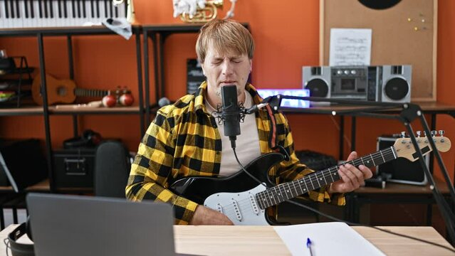 A young blond man with a beard plays guitar and sings into a microphone in a music studio, featuring equipment and instruments.