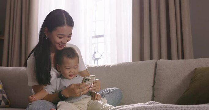 Asian mom and toddler spending time together. Happy smiling laughing multiethnic asian woman mom mother and son toddler baby kid child watching video using smartphone phone, laughing, enjoying time.