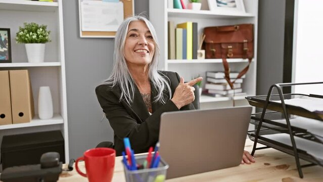 Middle age grey-haired woman using laptop working smiling happy pointing with hand and finger to the side at the office