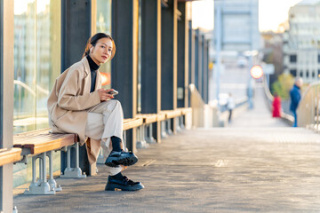 Naklejka premium Happy Asian woman using mobile phone with mobile app chatting or social media during waiting for tram at station. Attractive girl enjoy urban outdoor lifestyle travel city street with smartphone.