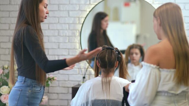 Female hairdresser mentor explains hair coloring procedure while newbie woman puts on black gloves standing behind female client in hair salon.