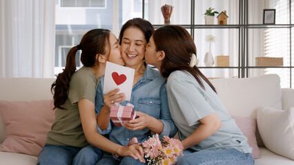 Mother day two grown up child cuddle hug give flower gift box red heart card to mature mum. Love kiss care mom asia middle age adult three people sitting at home sofa happy smile enjoy family time. - obrazy, fototapety, plakaty