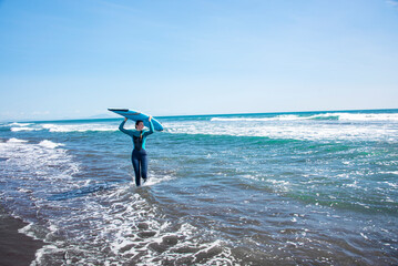There is a woman on the foreground. She is walking to the ocean  with surf board. She is a surfer. There is Pacific Ocean on the background.
