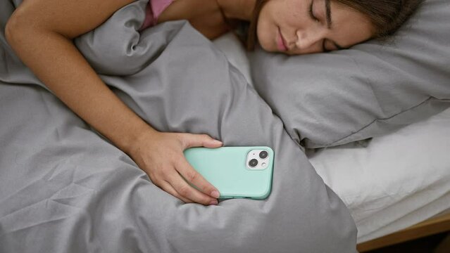 Sleepy young hispanic woman, comfortably lying in bed, holding her smartphone in a cozy bedroom - portrait of an attractive female relaxing at home, after a long day of exhaustion