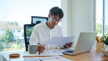 Fototapeta premium Asian businessman analyzing financial reports and using laptop at office.