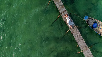Aerial view of a person relaxing on a rustic wooden pier over clear green waters next to a small...