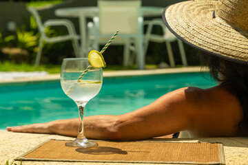 mulher na piscina encostada na borda usando chapéu de palha e bebendo um drink de limão com...