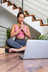 Vertical portrait of young Asian woman doing online meditation with hands in prayer sitting on yoga mat using laptop.