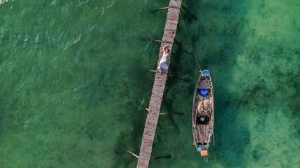 Aerial view of a person lying on a wooden pier over clear green waters next to a moored wooden boat