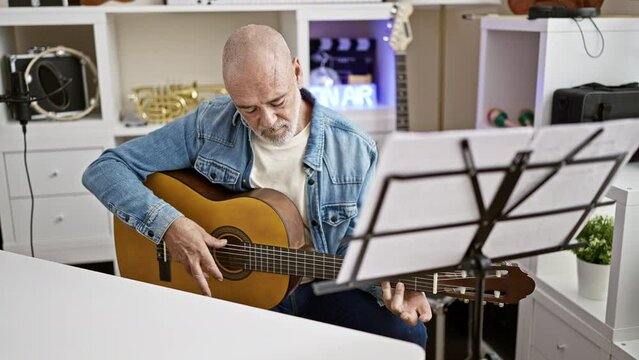 A focused senior man plays guitar in a home studio with musical instruments around him, practicing sheet music.