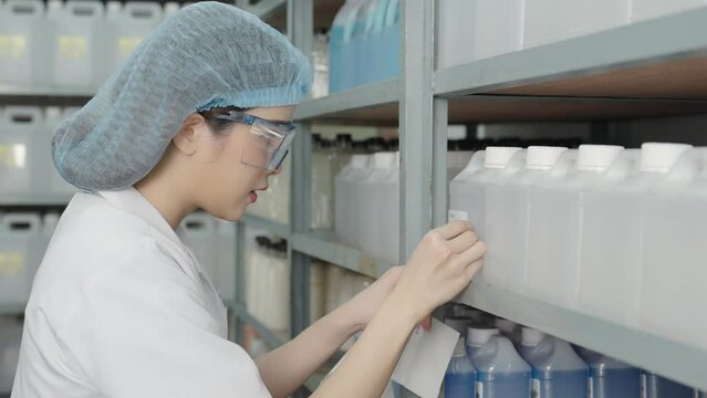 Beautiful young Asian chemist wears a hood, safety glasses puts a barcode sticker on a plastic bottle containing a solvent on a shelf in a store.
