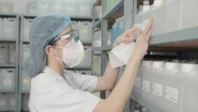 Beautiful young Asian chemist wears a hood, safety glasses, a face mask, puts a barcode sticker on a plastic bottle containing a solvent on a shelf in a store.