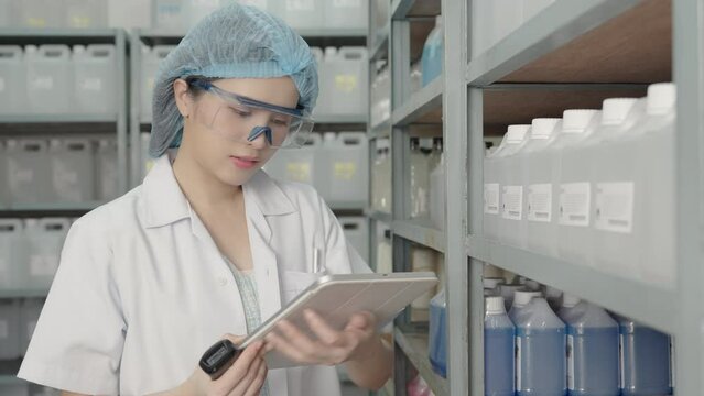 Beautiful young Asian chemist wears a hood, safety glasses using bar code reader to scanning bar code sticker on a plastic bottle containing a solvent on a shelf in a store to check information.