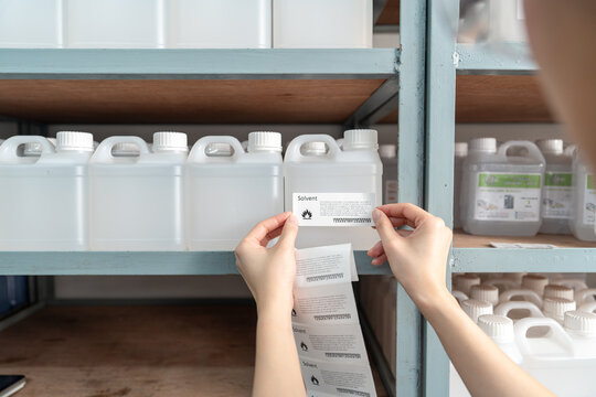 Beautiful young Asian chemist wears a hood, safety glasses puts a barcode sticker on a plastic bottle containing a solvent on a shelf in a stor