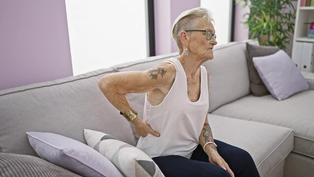 Unhappy grey-haired senior woman suffers severe backache, sitting on home sofa. worried, mature female adults grapple with serious spine problem indoors in middle age.