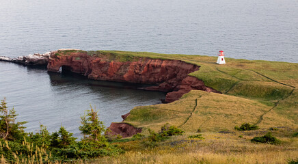 vue en hauteur d'un petit phare blanc et rouge avec du gazon vert en haut d'une falaise de roche rouge en bord de mer
