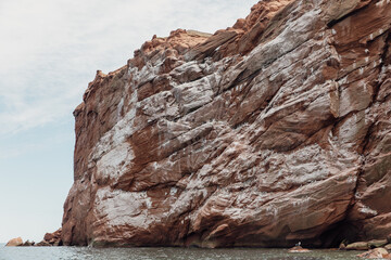 vue sur une haute falaise en roche rouge avec des fientes d'oiseau  au bord de l'eau en été lors d'une journée ensoleillée - obrazy, fototapety, plakaty
