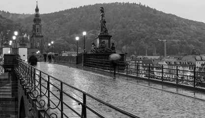Black and white photo of the old bridge (Karl-Theodor-Br&uuml;cke) in Heidelberg during a rainy day. Some pedestrians are walking with umbrellas. Blurred movement effect of the people on the bridge.