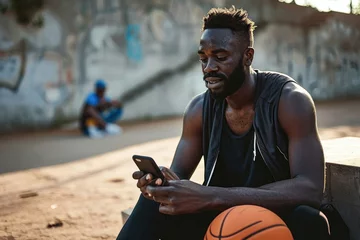 Foto op Canvas Fit sporty young African black ethnic man sitting outdoors holding basketball ball and mobile phone using apps, looking at smartphone, resting after street sport game, Generative AI  © Ashutosh