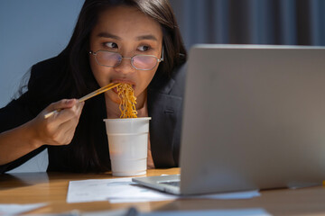 Asian business woman eating instant cup noodle while working on laptop in office at late night.