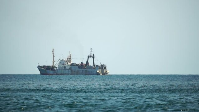 Fishing boat trawler catches fish while sailing on sea. A commercial fishing boat on the horizon in a distance sail to catch school of fish on calm sea surface in summer. Commercial catch of sea fish.