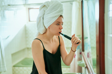 Young woman in a towel turban practices dental hygiene using an electric toothbrush in a home bathroom, focusing on teeth health with modern technology. - obrazy, fototapety, plakaty