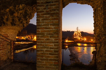 Magical Archway: A lit Christmas tree reflects in tranquil waters through a weathered brick arch....